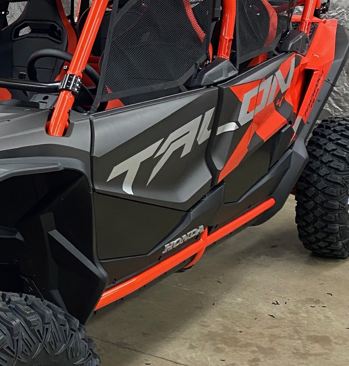 A close-up showcases an orange and black off-road vehicle with Talon branding, featuring large tires, a mesh seat cover, and rugged AJK Offroad Honda Talon Lower Doors made from CNC laser cut aluminum—ideal for tackling tough terrain.