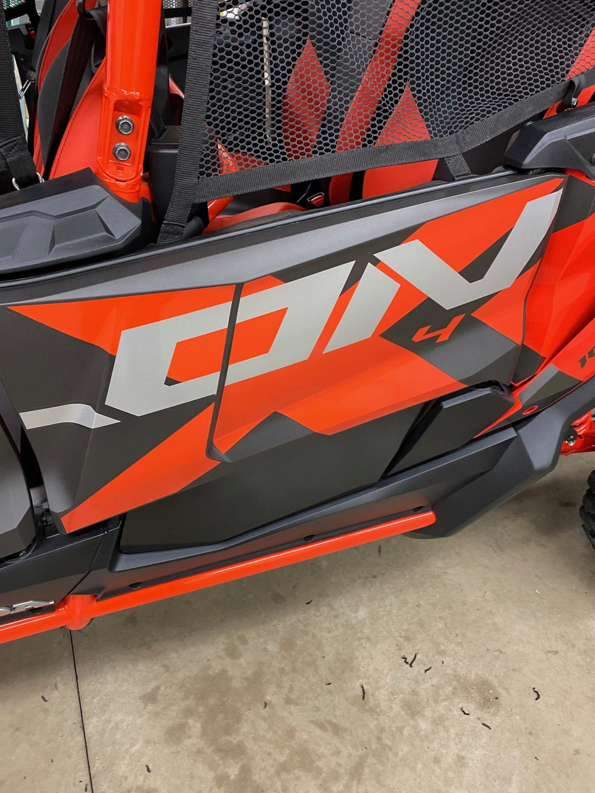 A close-up shows the red and black side panel of an off-road vehicle, designed with bold white text and geometric patterns, much like the AJK Offroad Honda Talon Lower Doors. The ground is concrete with scattered small debris.