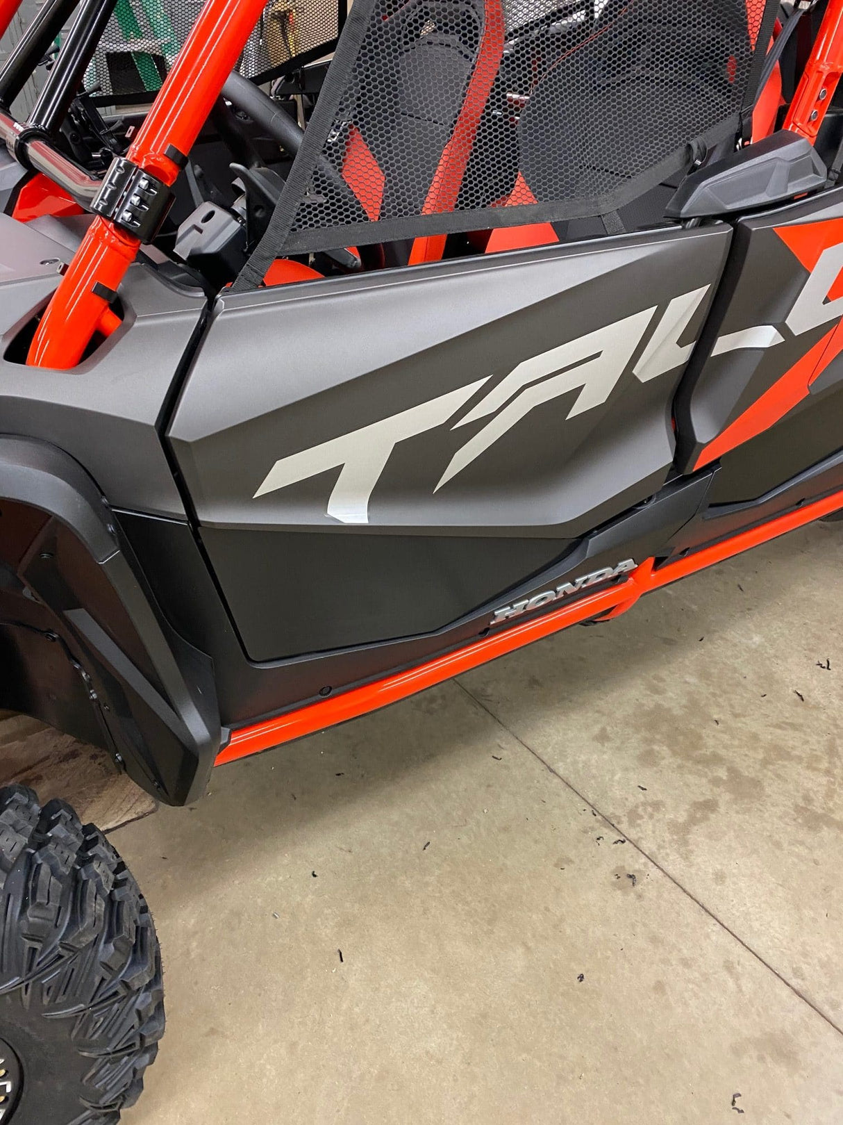 Close-up of a black and orange all-terrain vehicle with TALON on the side, featuring CNC laser-cut aluminum detailing from AJK Offroad. It has custom door bags, netted windows, visible large tires, and Honda Talon Lower Doors. Its parked on concrete.
