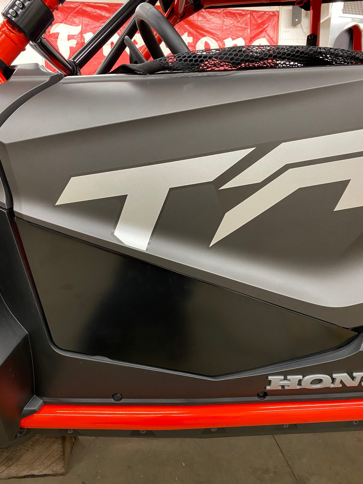 Close-up of a black and red Honda Talon Lower Doors panel by AJK Offroad, featuring sleek T4 and HONDA logos, crafted from CNC laser-cut aluminum. In the background, a garage and a red-and-white banner provide context.